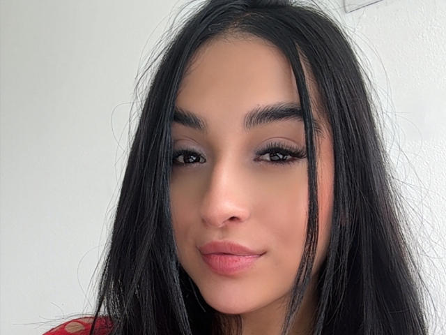 Close-up van EmmaWaltter, een jonge vrouw met lang zwart haar, volle lippen en donkere wenkbrauwen, die recht in de camera kijkt. camera met een lichte grijns. Witte achtergrond, lichte schaduw aan de rechterkant.