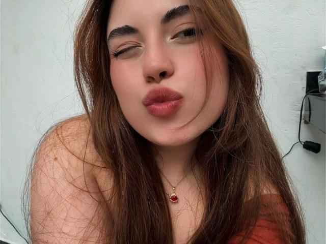 Close-up of VicluX with long brown hair, thick eyebrows, and red lipstick, puckering lips. VicluX has a light skin tone, wearing a red top, with a small gold necklace. Fucking sexy.