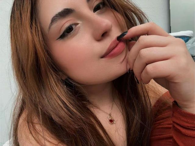 Primo piano di VicluX con capelli castano chiaro, che sorride sardonica, con eyeliner nero e smalto nero, mentre tocca le sue labbra. mano destra.
indossa una maglietta rossa e una collana sottile. Incredibilmente sexy..