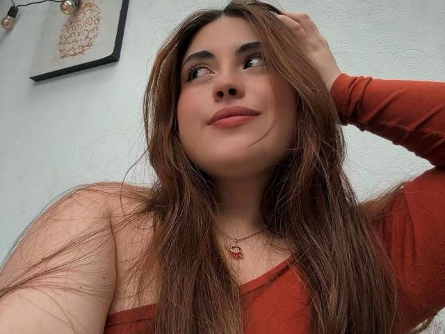 VicluX with long brown hair, wearing a red off-shoulder top, looking up with a slight smile, hand in hair, soft light, white wall, small framed art in background, subtle sexual tension.