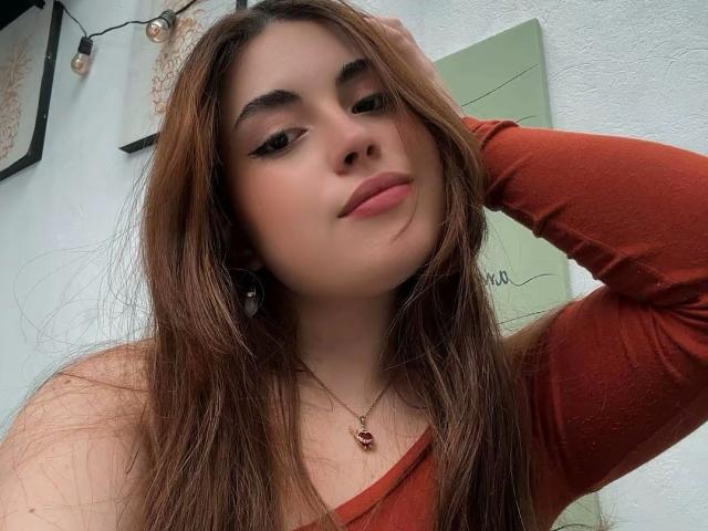 VicluX with long brown hair, wearing a red long-sleeve top, hand in hair, slightly tilted head, looking at camera, subtle smirk, necklace with small pendant, soft light, casual pose.