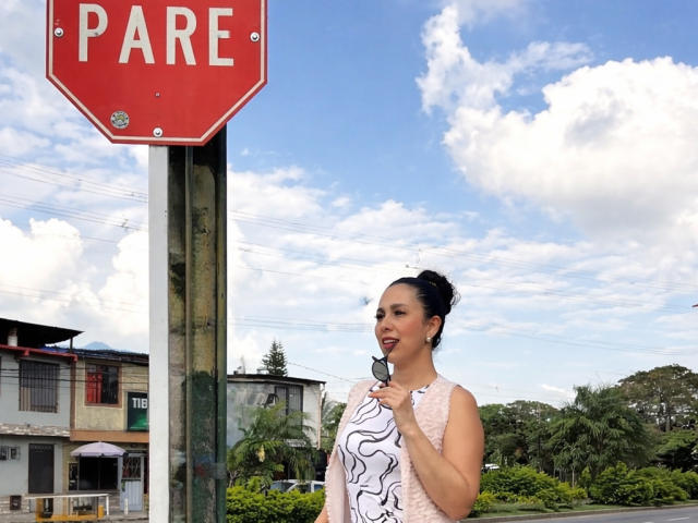 Katty Rivera staat naast een rood stopbord, ze draagt een wit tanktop met zwarte abstracte patronen en ze kijkt verleidelijk. naar de camera gericht. Blauwe lucht en wolken op de achtergrond. Warme, sensuele sfeer..