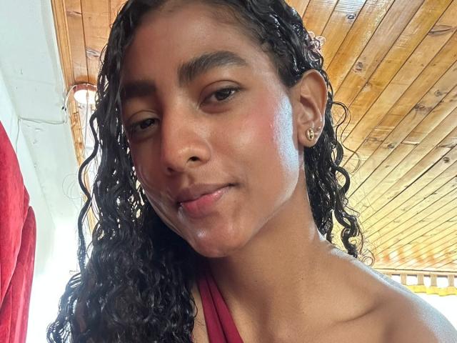 Close-up of NatalyBlonder, a tan-skinned woman with wet, curly black hair, wearing a red halter top, glistening skin, wooden ceiling background, slight smile, natural light, sexy, relaxed vibe.