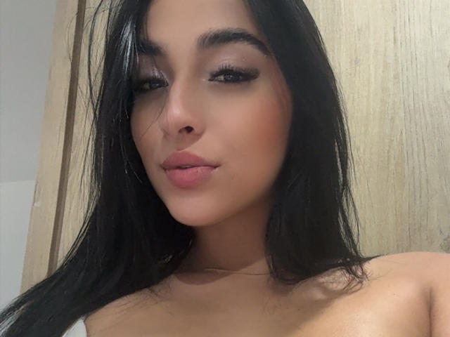 Primo piano di EmmaWaltter, una giovane donna con lunghi capelli neri, labbra piene e sopracciglia scure. Ha un sorriso appena accennato, le labbra leggermente dischiuse e un'espressione seducente. È senza top..