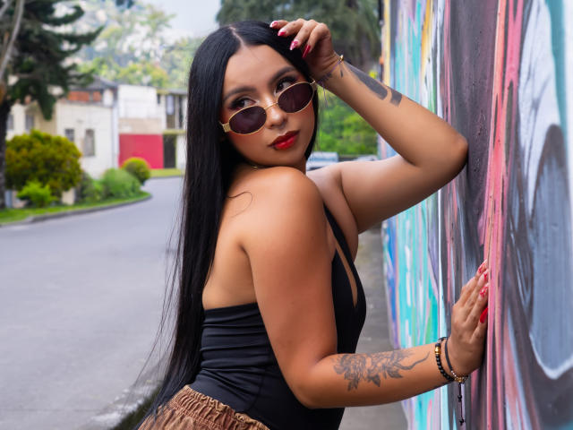 ChloeMoretti, uma mulher asiática de corpo curvilíneo com longos cabelos pretos, está encostada em uma parede colorida com grafites. Ela veste uma blusa preta sem alças, batom vermelho e óculos de sol redondos. A tatuagem no braço dela é visível. Incrivelmente sexy..