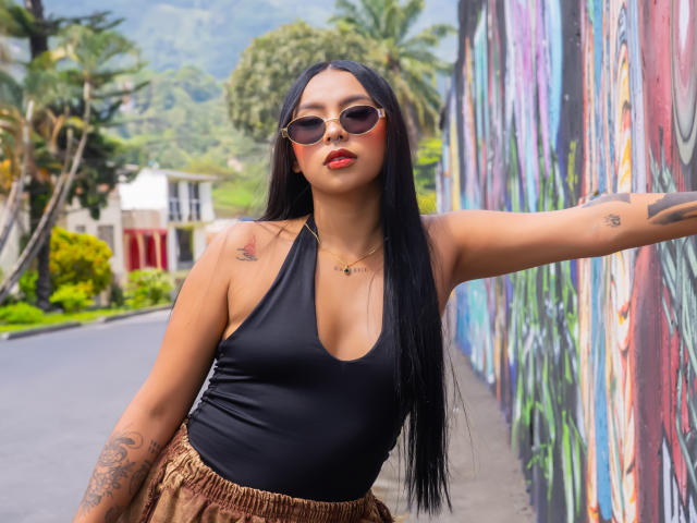 ChloeMoretti, une femme voluptueuse avec de longs cheveux noirs, portant un haut noir à encolure américaine et une jupe dorée, appuyée contre un mur coloré. mur avec des graffitis, tatouages sur son bras, lunettes de soleil, lèvres rouges, décor extérieur..