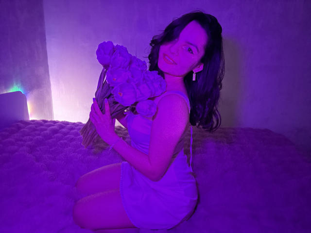 Photograph of JasmineAsha kneeling on a plush surface, holding a bouquet of flowers, bathed in purple and blue light. She's wearing a white dress, looking back with a smile. Her hair is wavy.