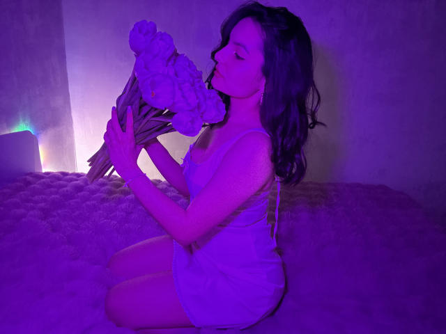 Photograph of a curvy woman with long dark hair, kneeling on a bed, holding a doll to her lips. Bathed in purple and blue light, her eyes are closed, exuding sexual tension.