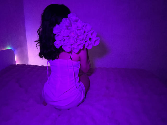JasmineAsha está de joelhos no chão, de costas para a câmera, vestindo um vestido branco com um grande arranjo de flores brancas. cabelo. Iluminado com luzes roxas e azuis. Tensão sexual..