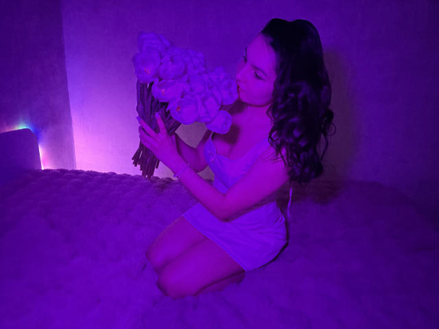 JasmineAsha kneels on a purple-lit carpet, holding a bouquet of white flowers. She's topless, with curly black hair, and eyes closed, creating a sensual, intimate vibe. Her pose is submissive.