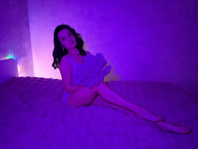 JasmineAsha, una mujer de cabello oscuro, sentada sobre una superficie esponjosa, vestida con un vestido blanco de encaje, sosteniendo un ramo de flores. bañada en luz púrpura y azul, con las piernas cruzadas, en una pose seductora..