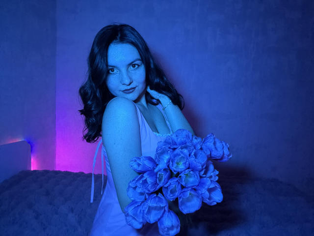 JasmineAsha, fotografía con iluminación azul, sosteniendo un ramo de flores azules grandes, mirada seductora, cabello oscuro ondulado, vistiendo una vestido blanco de encaje, fondo con luces moradas y azules, tensión sexual..