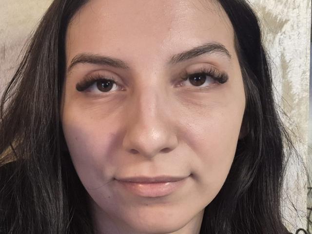 Close-up of SeleneNorth, a young woman with long dark hair, fair skin, and thick eyebrows. She has subtle makeup, full lips, and brown eyes. Sexual tension evident in her slight smile.