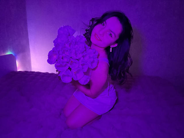 JasmineAsha, kneeling on a carpet, holding a large bouquet of white flowers, illuminated by purple and blue lighting. She has long, wavy dark hair, wearing a white dress. Sexual tension.