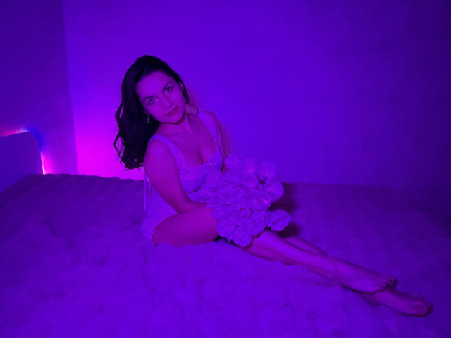 JasmineAsha, with long black hair, sits on a bed under purple and pink lighting. She wears a white, floral, lace dress, legs crossed, leaning slightly forward, seductive gaze.