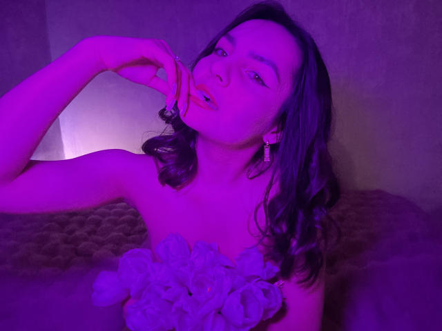 Fotografia di JasmineAsha con illuminazione viola, capelli ondulati scuri e un'espressione seducente. Lei si sta mordendo il dito, indossa una camicetta bianca con volant, con un décolleté evidente e uno sguardo malizioso..