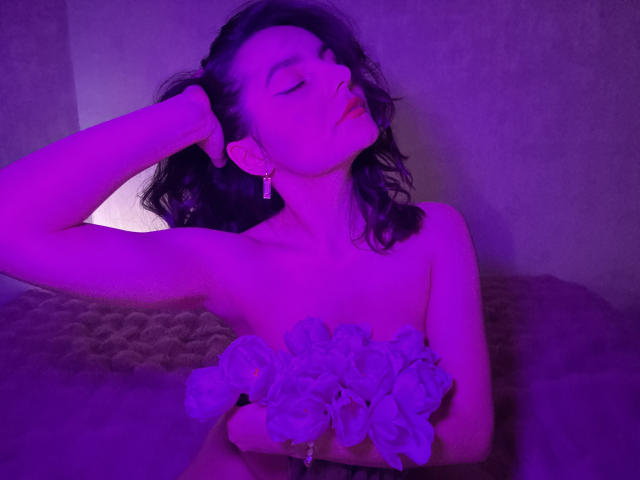 JasmineAsha with closed eyes and head tilted back, holding a bouquet of white flowers, bathed in purple light. Her right arm is raised, hand behind her head, exuding sexual tension.