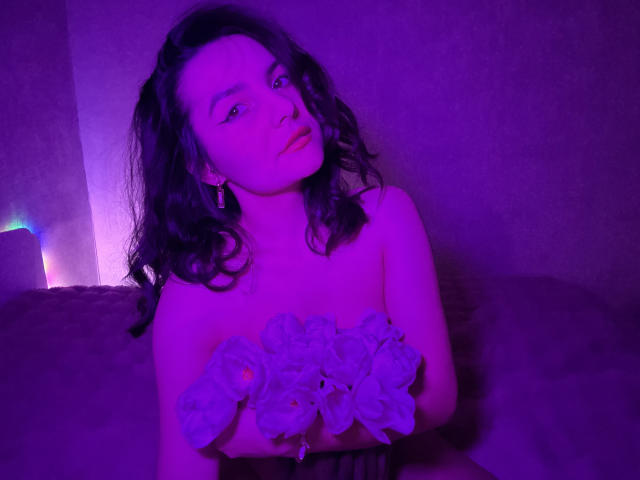 Photograph of JasmineAsha with shoulder-length curly hair, illuminated by purple and blue lighting. She holds a bouquet of flowers, topless, looking up with a seductive expression, cock partially visible.