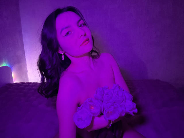 Photograph of JasmineAsha with purple and blue lighting, sitting on a couch, holding a bouquet of white flowers, looking up with a slightly open mouth, wearing a strapless dress.