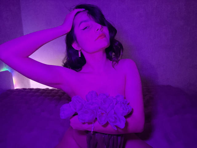 Photograph of JasmineAsha with dark, wavy hair, eyes closed, hand in hair, wearing a strapless dress with ruffles, bathed in purple and blue light, looking seductive and relaxed.