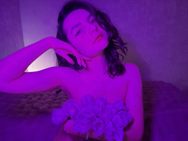 Fotografía de JasmineAsha con iluminación púrpura, recostada con un brazo levantado, sosteniendo un trozo de tela blanca con volantes. pecho. Ella tiene el pelo negro y ondulado, los ojos entrecerrados y una expresión sensual..