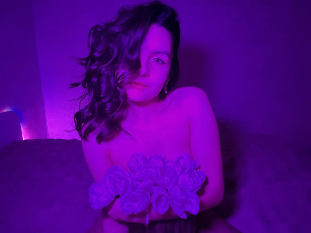 Photograph of JasmineAsha, illuminated by purple and blue lighting. She has wavy black hair, partially covering her face, wearing a strapless top with floral patterns. She's looking seductively at the camera.