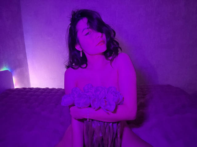 Photograph of JasmineAsha with wavy black hair, sitting on a couch under purple and blue lighting. She's wearing a sheer, ruffled top, with a sultry, seductive expression. Her hands are on her thighs.