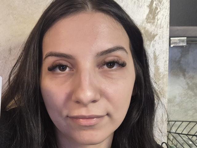 Primo piano di SeleneNorth, una giovane donna con lunghi capelli neri, pelle chiara e occhi marroni, che guarda direttamente verso la fotocamera. con un leggero sorriso. Lo sfondo include una parete bianca e una ringhiera in metallo. La sua espressione trasmette una sottile tensione sessuale..