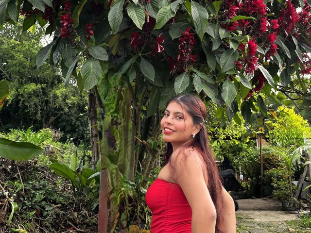 AlyssaGreen with long brown hair stands under a tree with red flowers, wearing a tight red strapless top, smiling at the camera. Dense green foliage in the background. Sexual tension.