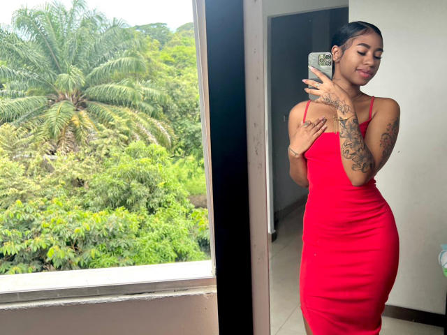 Photographie de LovelyPeachss debout dans un encadrement de porte, prenant un selfie dans un miroir. Elle porte une robe rouge moulante, qui révèle des tatouages sur son bras et son côté. Végétation tropicale à l'extérieur. Putain, c'est sexy..