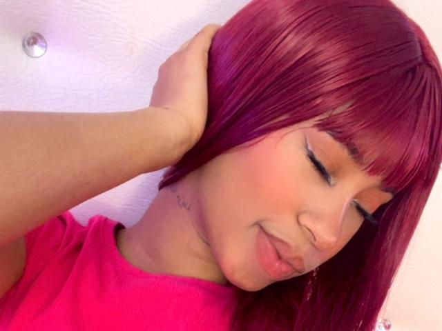 Close-up foto van LovelyPeachss met recht, felrood haar, een pony en een roze top. Ze legt haar hand op haar hoofd, haar ogen zijn gesloten, haar lippen zijn lichtjes samengeknepen, en ze straalt een subtiele seksuele spanning uit. Helder licht.