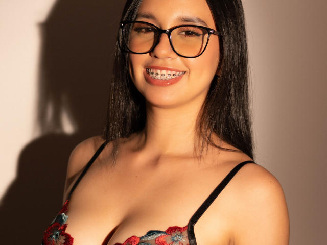 Close-up photo of Keylamml, an Asian woman with long black hair, wearing black-framed glasses and a red lace bra, smiling with braces. Strong sunlight casts shadows on her face.