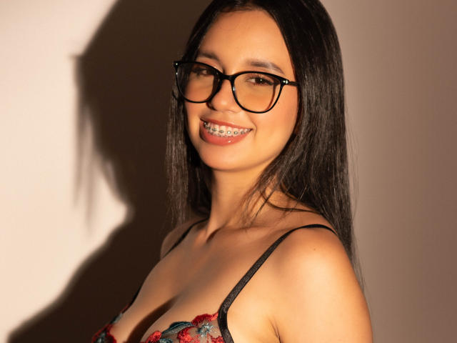Photograph of Keylamml, a smiling woman with long black hair, wearing black-framed glasses and a red floral bra, showcasing her cleavage, with shadowed light highlighting her sexy grin.