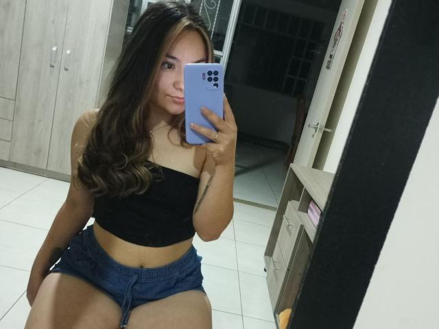 Elizabeth White, met lang, golvend bruin haar, draagt een zwarte top zonder bandjes en blauwe shorts, en houdt een blauwe telefoon voor zich. van een spiegel, zittend, een spiegel-selfie maken, seksuele spanning.