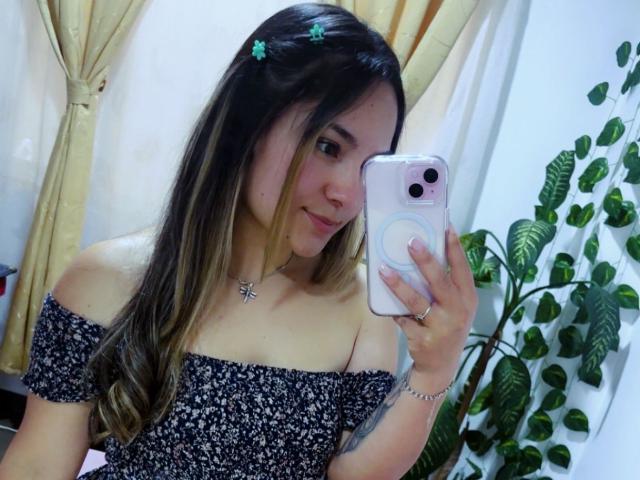 Elizabeth White, met lang bruin haar, draagt een zwarte bloemenjurk met een open schouderlijn en maakt een spiegelzelfportret met een witte iPhone. Ze heeft groene haarspelden, een ketting en een ring. Super schattig..