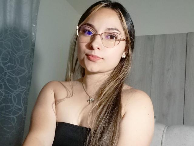 Fotografía de Elizabeth White con cabello largo y castaño, usando gafas y un top negro sin tirantes, mirando directamente a la cámara. Tiene una ligera sonrisa, piel clara y un collar de plata. Su expresión es increíblemente segura..