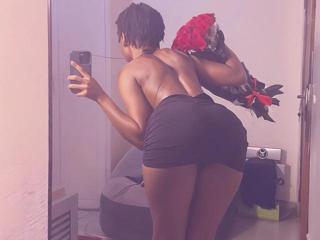 Una mujer atractiva de piel oscura, con cabello negro corto, sin ropa en la parte superior, inclinada, sosteniendo rosas rojas, vestida con pantalones cortos negros, tomándose una foto en un espejo. selfie, mostrando un trasero curvilíneo, teléfono en la mano izquierda, habitación blanca, luz suave..