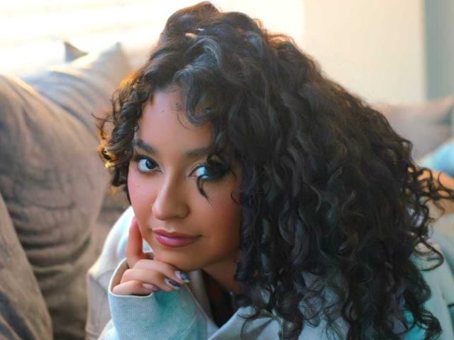 Photo en gros plan de CarmilleZlata, une femme voluptueuse aux cheveux noirs et bouclés, appuyant son menton sur sa main, avec un regard séduisant. Sur le côté, elle portait un sweat à capuche gris clair et avait un maquillage éclatant..