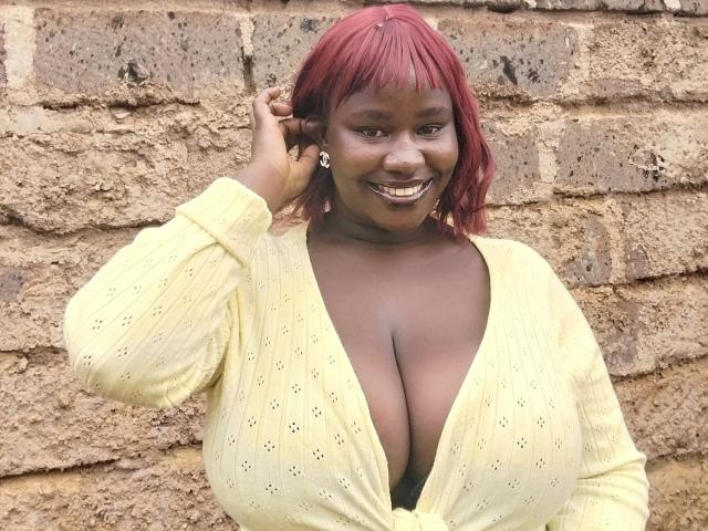 CathrynStella with red hair, smiling, wearing a yellow, deep-cut, knit top, showing large, round, bare breasts against a rough brick wall.