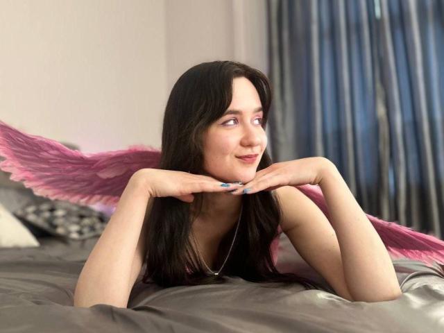 Œuvre d'art numérique de BerryDiva, avec de longs cheveux noirs, allongée sur un lit avec des ailes roses en plumes, les mains sous le menton, souriante. De manière séduisante, éclairage doux, rideaux bleus en arrière-plan, tension sexuelle..