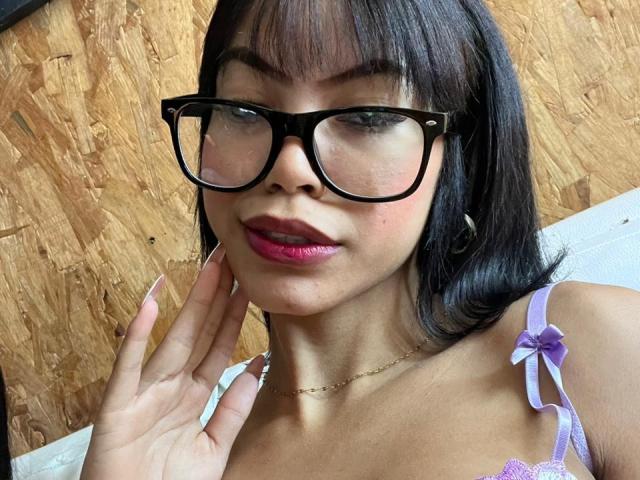 Close-up de MaliaFoxxy usando óculos de aro preto, com cabelo preto e liso, e batom roxo. Ela está usando um sutiã de renda roxo e tocando sua bochecha com a mão. Fundo de madeira. Olhar sensual e confiante..