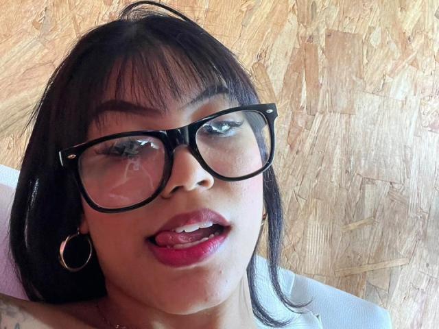 Close-up of MaliaFoxxy, a young woman with black hair and bangs, wearing large black glasses, red lipstick, and hoop earrings, sticking her tongue out suggestively. Light wood background.