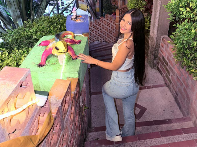 EstrellaXHot, curvy, with long black hair, wearing a white crop top and blue jeans, leans on a brick wall, looking back, next to a yellow and red toy on a green surface.