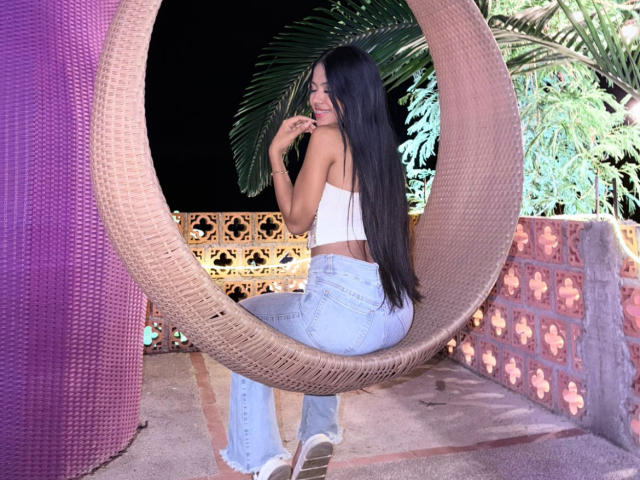 EstrellaXHot, cabello largo y negro, camiseta blanca, jeans azules, sentada en una silla grande de mimbre, con el atributo sexual visible, pose seductora. mano cerca de la boca, plantas tropicales, pilar púrpura, ambiente nocturno.