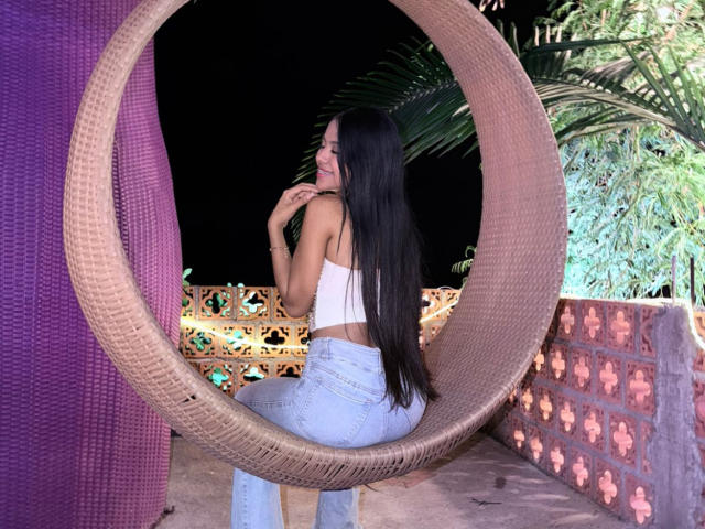 EstrellaXHot, con cabello largo y negro, vestida con un top corto blanco y pantalones blancos ajustados, está sentada en un gran asiento circular de mimbre. Encuadre, mirando hacia atrás con una mano cerca de la boca. Entorno exterior vibrante.