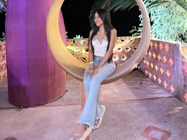 EstrellaXHot est assise sur une balançoire en métal de couleur violette, vêtue d'un haut blanc sans bretelles et d'un jean bleu clair usé. Ses longs cheveux noirs tombent sur ses épaules. Elle regarde vers le bas, avec une posture détendue..