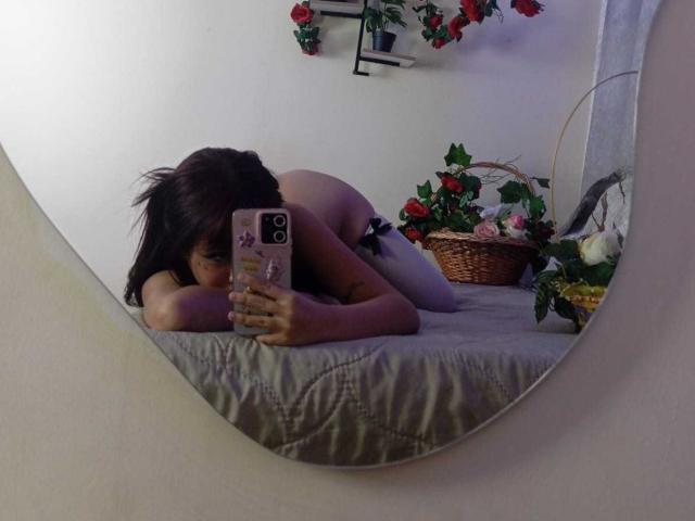 KatheLux lies nude on a bed, face down, taking a mirror selfie with a phone. She has dark hair covering her face. A basket with red roses is in the background. Fucking sexy.