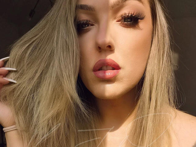 Primer plano de KimJudge, atractiva, con cabello rubio, labios carnosos con un brillo labial rosa, delineador de ojos negro intenso, uñas largas y blancas. labios ligeramente entreabiertos, rostro iluminado por el sol, fondo oscuro, mirada intensa, ambiente sensual..