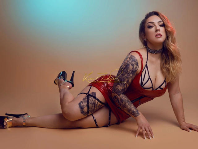 KimJudge, de aspecto atractivo y con numerosos tatuajes, lleva ropa interior roja con tirantes y tacones altos negros, y está acostada en un suelo color arena, con un brazo apoyándola. pierna doblada, pene visible a través de la ropa interior, mirada seductora, iluminación de estudio..