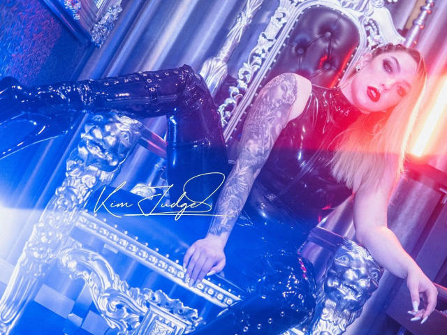 KimJudge – heiß, unglaublich sexy, lehnt sich in einem Stuhl zurück, mit einem leuchtenden roten Licht in ihrem blonden Haar, trägt ein glänzendes schwarzes kleid aus Latex, eine blau leuchtende Gitarre haltend..